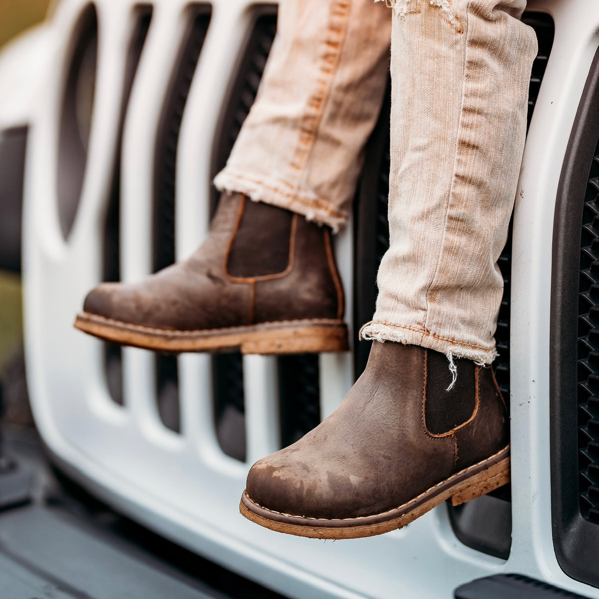 Chocolate Chelsea boots worn with light jeans on the front of a white Jeep.