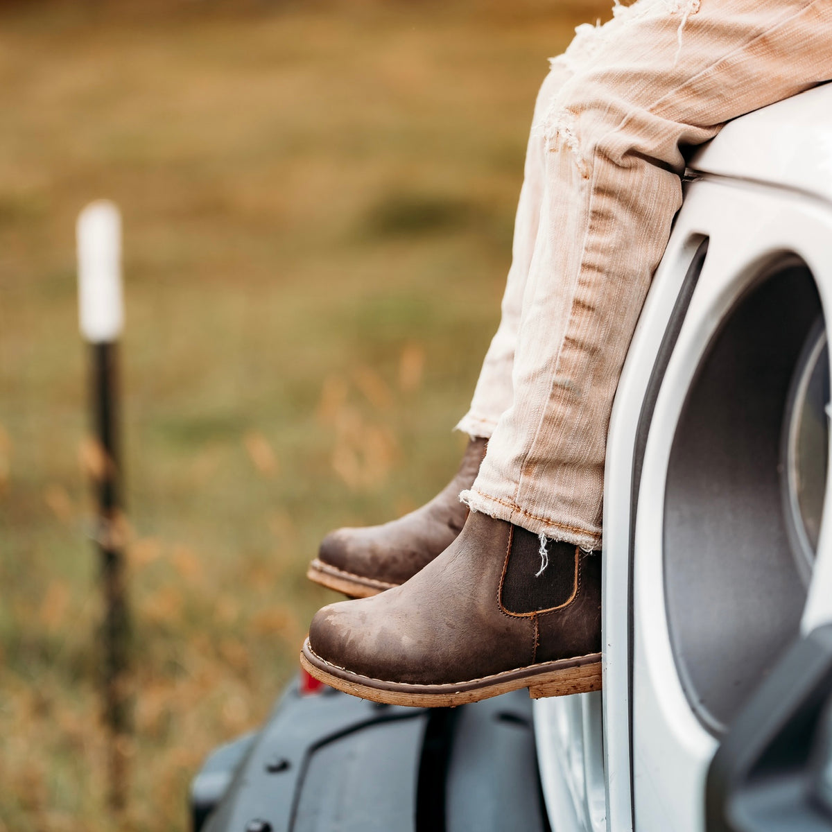 Chocolate Chelsea boots and light jeans beside a white Jeep outdoors.