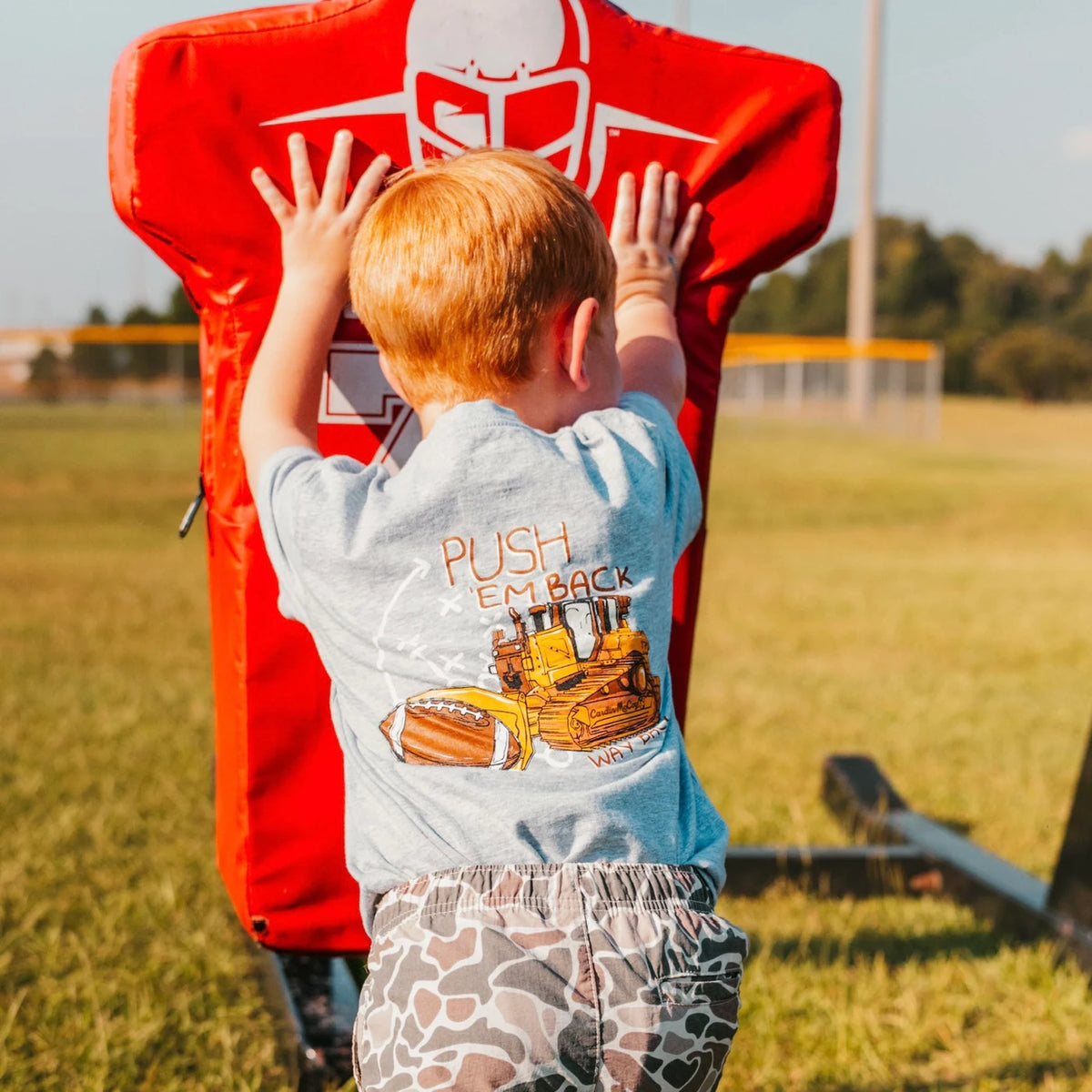 A child wearing a Push Em Back football tee while pushing a tackling dummy.