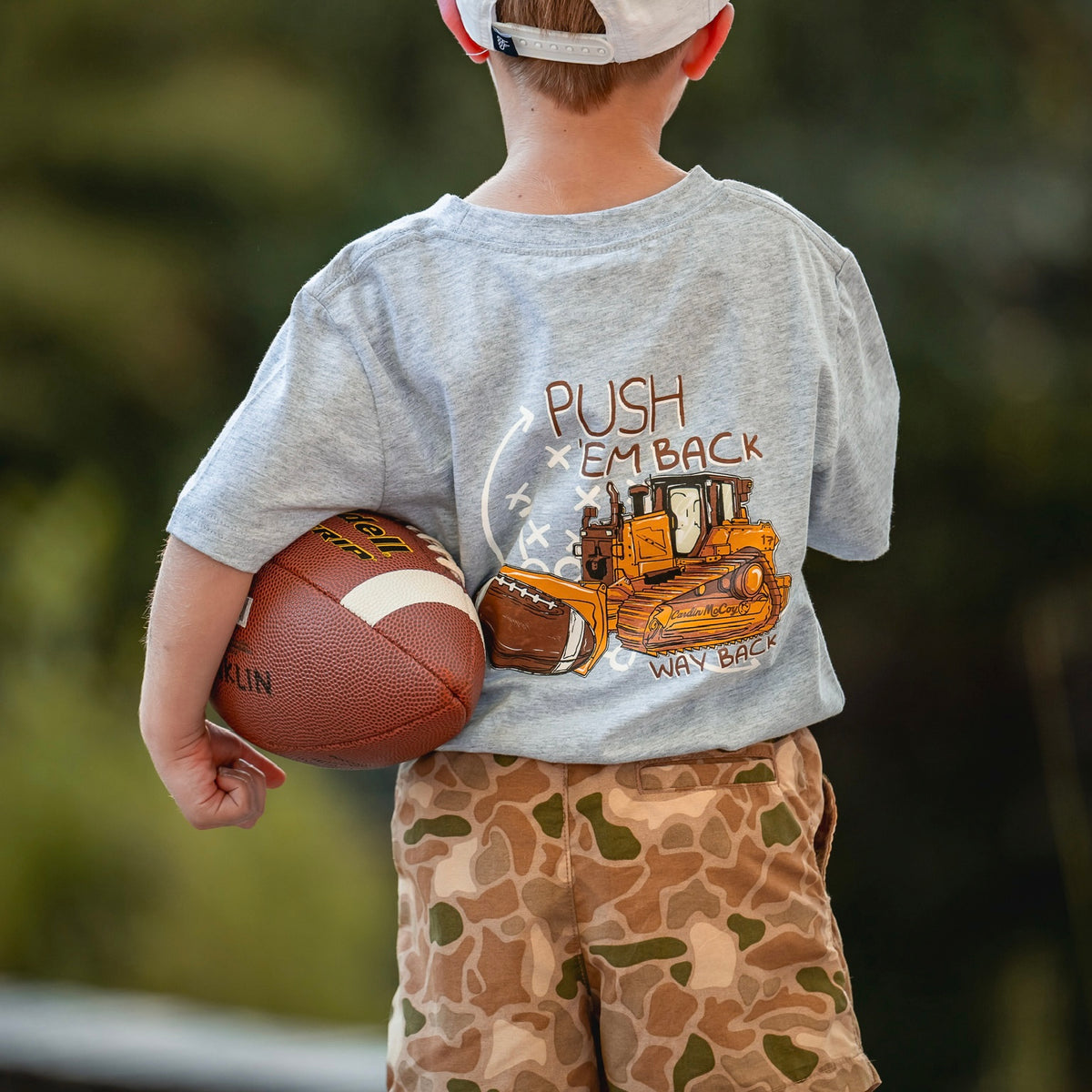 A child holding a football in a football graphic tee and camo shorts.