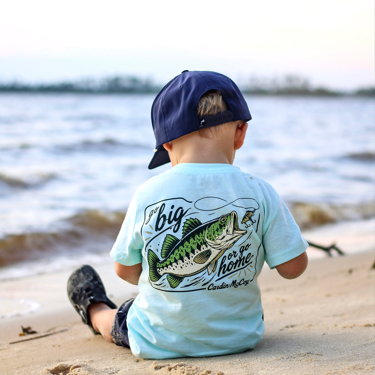 A child wearing a fishing graphic tee beside the water.